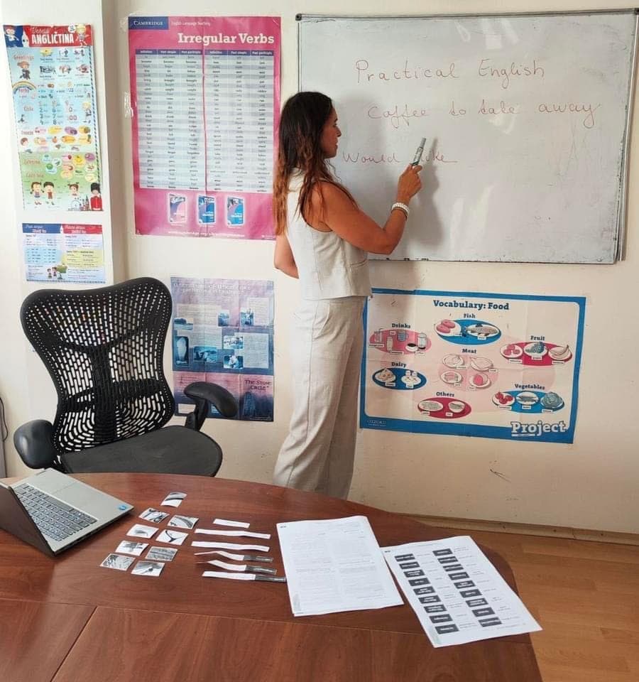 A classroom: someone writing on the whiteboard, grammar and vocabulary charts on the walls.